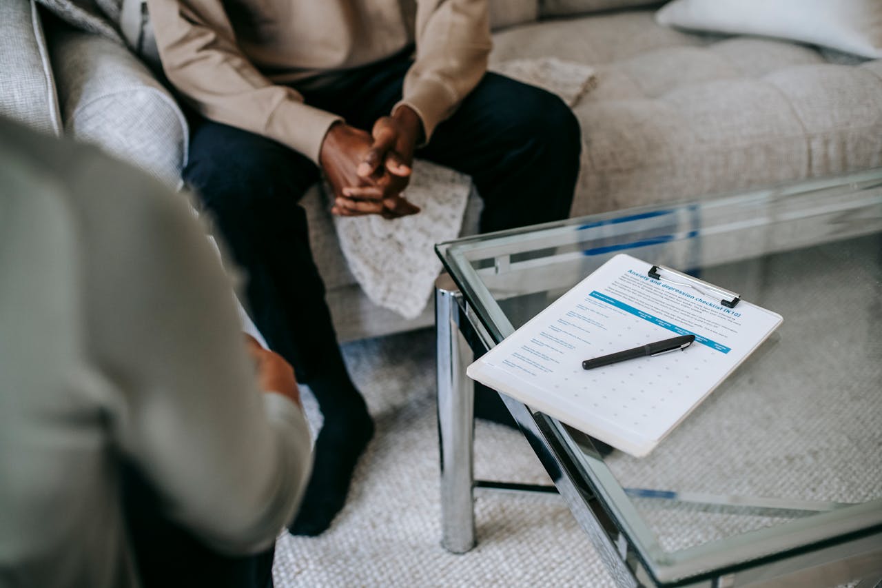 From above of unrecognizable ethnic male patient with clasped hands sitting near anonymous psychologist in office with glass table and clipboard with notes